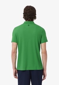 Uomo con capelli ricci castani che indossa una polo a maniche corte di un vivace verde e pantaloni blu scuro, in piedi con la schiena rivolta in avanti.