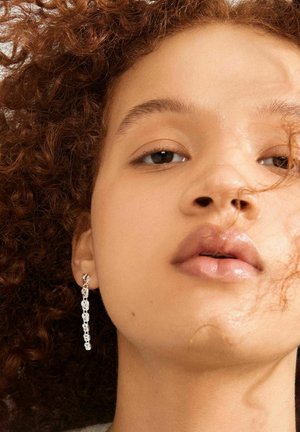 Close-up of a person with curly hair wearing a long, silver chain-link earring, with a neutral expression and natural makeup.