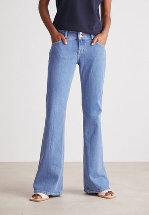 Osoba nosząca jasnoniebieskie dzwony z wysokim stanem i podwójnymi guzikami, z rękami w kieszeniach, granatową bluzkę i białe sandały z odkrytymi palcami.