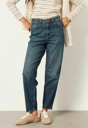 Frau trägt blaue High-Waist-Jeans, beige Sneakers, ein gestreiftes langärmliges Shirt und einen cremefarbenen Strickcardigan, der über die Schultern gelegt ist, Hände in die Hüften gestützt.