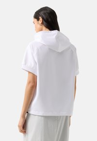 Mujer con cabello largo y oscuro, vistiendo una sudadera blanca de manga corta y suelta y una falda de color claro, de espaldas contra un fondo liso.
