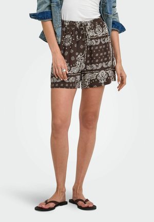Kvinde iført brune mønstrede shorts med snøre, hvid top, denimjakke og sorte flip-flops, der står mod ensfarvet baggrund.