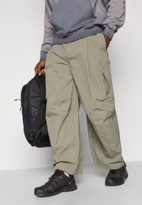 Calças cargo largas, de tom cáqui claro, com bolsos com fecho, combinadas com um sweatshirt cinzento e sapatos de caminhada pretos, segurando uma mochila preta.