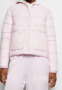 Persona che indossa una giacca a vento rosa chiaro con bottoni a pressione e grandi tasche frontali, abbinata a pantaloni rosa chiaro coordinati.