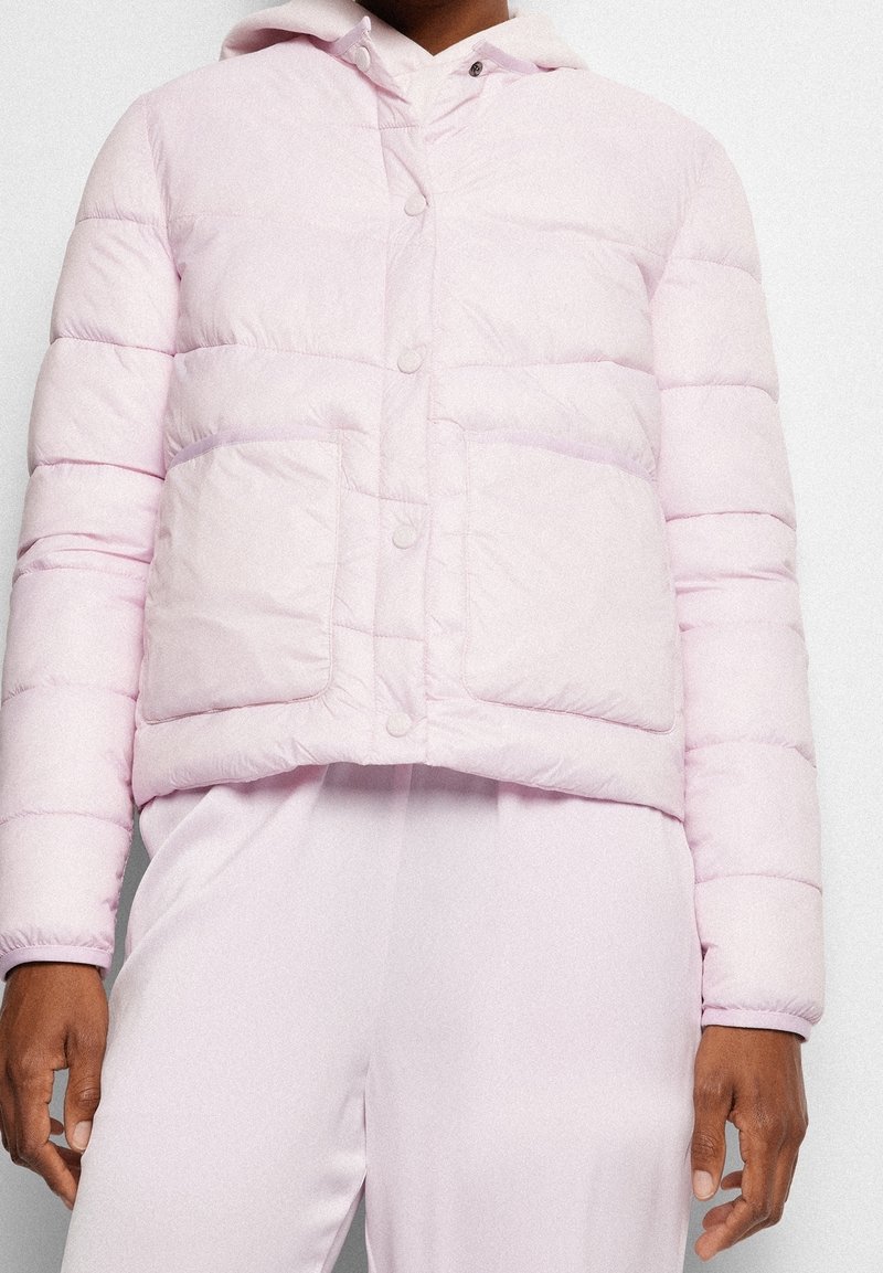 Persona che indossa una giacca a vento rosa chiaro con bottoni a pressione e grandi tasche frontali, abbinata a pantaloni rosa chiaro coordinati.