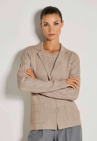 Frau mit dunklen Haaren, die zurückgebunden sind, trägt einen beigefarbenen Strickcardigan über einem passenden Pullover und grauen Hosen, steht mit verschränkten Armen vor einer weißen Wand.