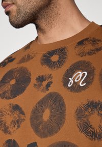 Καφέ T-shirt με μαύρο κυκλικό γραφικό σχέδιο, διαθέτοντας υφασμάτινη υφή και κεντημένο λευκό λογότυπο στο στήθος.