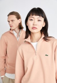 Due modelli indossano felpe rosa chiaro con zip, dotate di un piccolo logo verde a forma di coccodrillo sul lato sinistro del petto. Il tessuto sembra morbido e liscio.