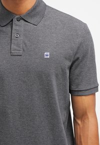 Ciemnoszara koszulka polo z bawełny z kołnierzykiem, krótkimi rękawami, dwoma guzikami i małym niebieskim logo na piersi. Materiał o fakturowanej strukturze.