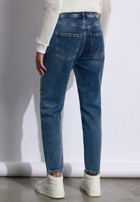 Vaqueros de mezclilla en azul medio con un corte recto, dos bolsillos traseros y un ligero desgastado. Combinados con zapatillas blancas.