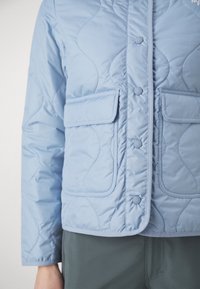 Jasnoniebieska pikowana kurtka z dwoma przednimi kieszeniami z klapkami, zapięciem na napy i gładką fakturą. Charakteryzuje się subtelnym falowanym przeszyciem.