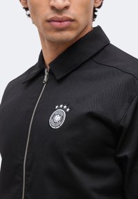 Κοντινό πλάνο ενός ατόμου που φοράει μαύρο μπουφάν με φερμουάρ, με το λογότυπο της Deutsche Fußball-Bund και τέσσερα αστέρια στο αριστερό στήθος.