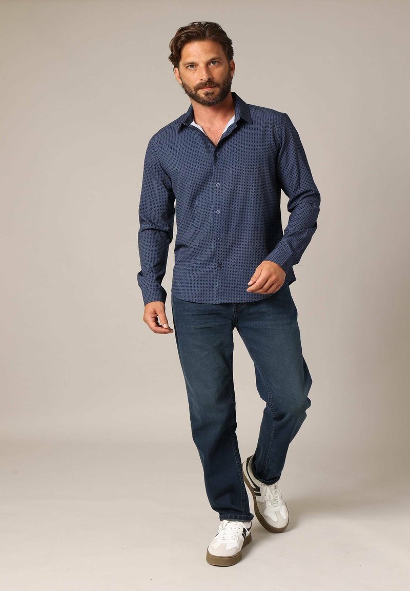 Marineblauwe overhemd met kleine blauwe stippen, lange mouwen en een witte kraag aan de binnenkant, gecombineerd met donkere jeans en witte sneakers.