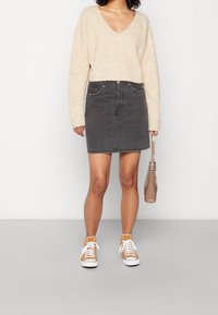 Pull beige côtelé avec un col en V, associé à une mini-jupe en jean grise. Porté avec des baskets montantes orange et tenant un petit sac à main marron.