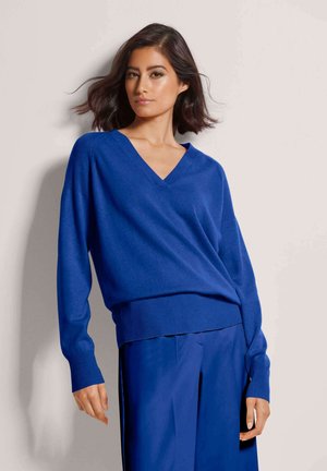 Vrouw met donker haar tot op schouderlengte, die een blauwe trui met V-hals en bijpassende blauwe broek draagt, staand tegen een effen lichte achtergrond.