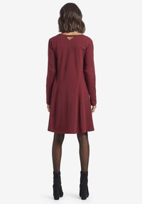 Abito lungo bordeaux a maniche lunghe con orlo svasato, una scollatura a chiave sul retro e tessuto strutturato. Indossato con stivali neri alla caviglia e collant.