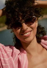 Lunettes en écaille de tortue avec des verres sombres, associées à une chemise à rayures roses. Des cheveux bouclés encadrent le visage, réfléchissant la lumière du soleil.