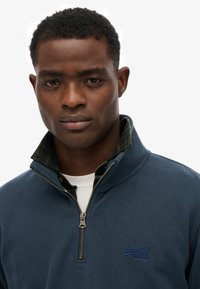 Granatowy sweatshirt na zamek z teksturowanej tkaniny, zapinany na kołnierz, z niebieskim logo. Nałożony na zieloną koszulę w kratę. Posiada srebrny zamek.