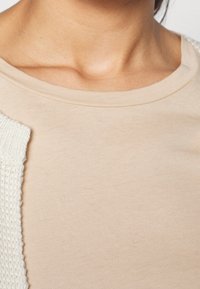 Beżowa bawełniana koszulka z okrągłym dekoltem. Posiada teksturowany biały akcent na jednym ramieniu. Gładka powierzchnia materiału.