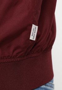 Cazadora bomber burdeos con cintura elástica, textura suave y etiqueta de tela blanca que muestra "JACK & JONES EST 1990" en el lado.