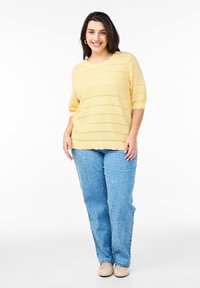 Haut en tricot jaune à manches courtes avec des motifs en zigzag, associé à un jean texturé bleu clair. La silhouette se tient sur un fond blanc uni.
