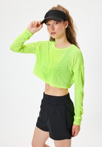 Neon grøn cropped langærmet top med tekstureret stof og shorts med sort elastisk taljebånd. Modellen bærer en sort visir.