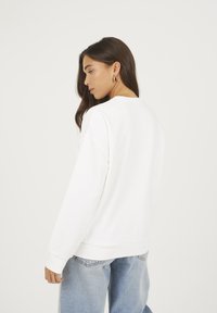 Giovane donna con i capelli lunghi e scuri indossa orecchini a cerchio dorati, una felpa bianca e jeans azzurri chiari, girandosi leggermente di lato.