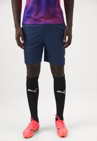 Camisa de manga curta em gradiente roxo e rosa, calções azul-marinho, meias de joelho pretas com logótipo branco e sapatilhas de futebol cor-de-rosa brilhante.