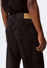 Black denim jeans feature a straight-leg design, five pockets, and a tan leather label with the word "WEEKDAY" on the back waistband.