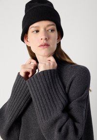 Pull en maille côtelée gris foncé avec un col montant, associé à un bonnet en tricot noir texturé. Matière douce visible dans les détails et les plis du tissu.