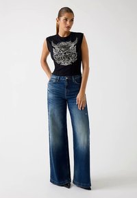 Top nero senza maniche con design grafico abbinato a jeans denim blu ampi, leggermente usurati. Tessuto liscio, vestibilità morbida, lavaggio medio.