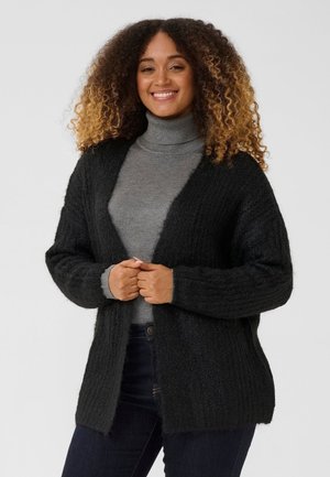 Une femme souriante porte un cardigan noir en maille par-dessus un col roulé gris et un jean foncé, sur un fond clair et uni.