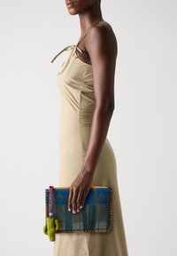 Farbenfrohe Mesh-Clutch mit einem Kaktus-Akzent, die eine Mischung aus Blau, Grün und Orange zeigt. Flaches Design, gehalten von einer Person in einem beigen Kleid.