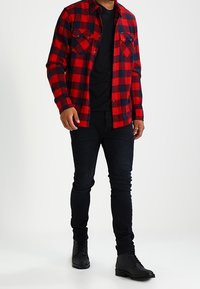 Camisa de franela a cuadros rojos y azul marino con dos bolsillos frontales, llevada sobre una camiseta negra, combinada con unos jeans ajustados negros y botas negras.