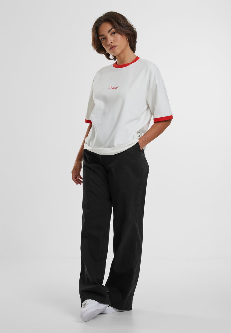 T-shirt bianco a maniche corte con colletto e polsini rossi, caratterizzato da un piccolo logo rosso. Abbinato a pantaloni neri larghi e sneakers bianche.