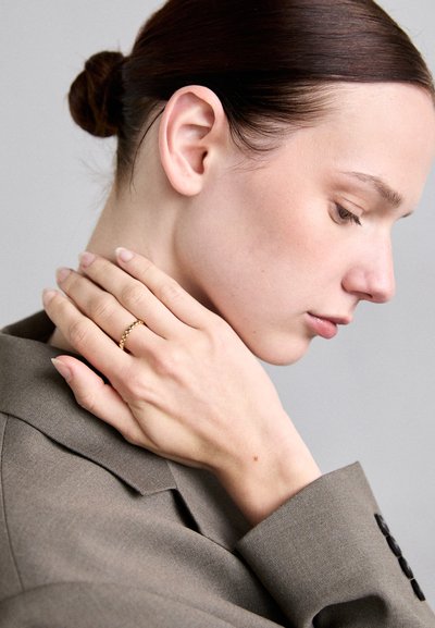 Goldring mit einem verknüpften Design an einer Hand, vor einem neutralen Hintergrund. Das Modell trägt ein maßgeschneidertes braunes Blazer und hat einen tiefen Dutt.
