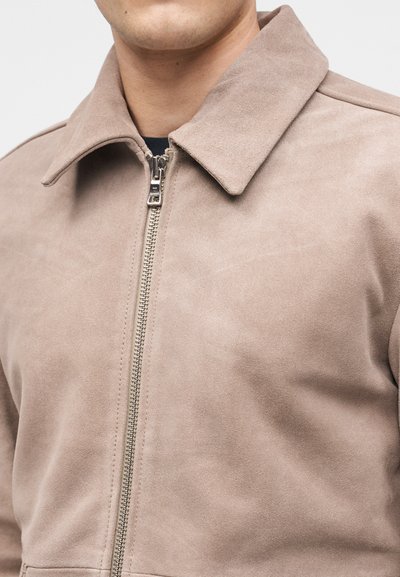 Άνδρας που φοράει μπεζ σουέτ μπουφάν με ασημί φερμουάρ μπροστά και μυτερό γιακά, εμφανίζεται από το στήθος και πάνω.