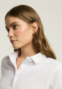Biała koszulka polo z teksturowanej tkaniny, z płaskim kołnierzem i trzema guzikami przy szyi. Modelka ma długie brązowe włosy i naturalny makijaż.