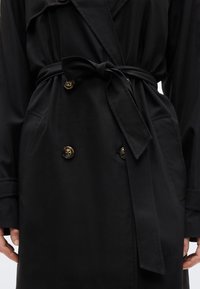 Κοντινό πλάνο μαύρου παλτού τύπου trench με κουμπιά από τορτοϊσέλ και δεμένη ζώνη στη μέση, εμφανίζοντας τσέπες και μακριά μανίκια.