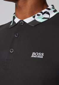 Černé polo tričko s ohraničením vzorem v mátově zelené a černé barvě. Obsahuje tři knoflíky a vyšitý logo v bílé barvě na hrudi.