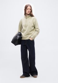 Γυναίκα με μπεζ φούτερ με φερμουάρ ως το στήθος, ευρύχωλο σκουρόχρωμο τζιν, κρατώντας μαύρο δερμάτινο μπουφάν, στέκεται μπροστά σε λευκό φόντο, κοιτάζοντας προς τα αριστερά.