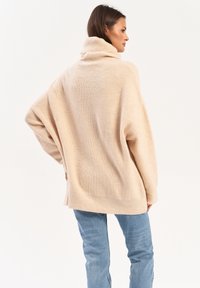Oversized beżowy sweter z wysokim, złożonym kołnierzem i prążkowaną fakturą. Posiada opadające ramiona i boczne rozcięcia dla swobody ruchów.
