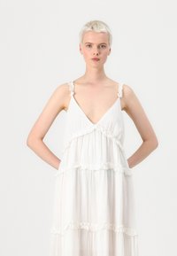 Hvit lagdelt kjole med volanger på stroppene og V-utringning. Lett, flytende stoff med horisontale volanger over hele kjolen. Enkel design.