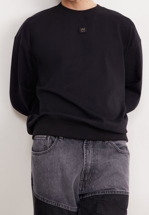 Person iført en sort langærmet sweatshirt med et lille logopatch på brystet og tofarvede sorte og grå jeans.