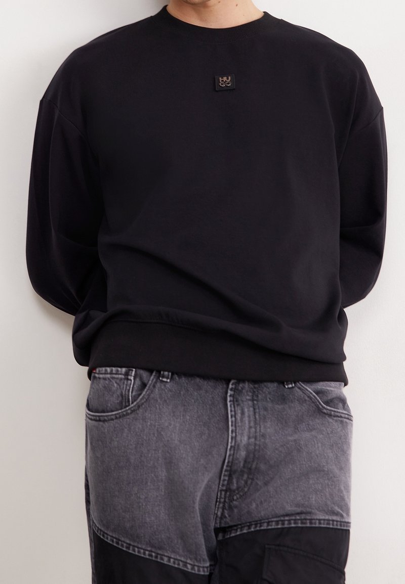 Personne portant un sweat-shirt noir à manches longues avec un petit écusson logo sur la poitrine et un jean bicolore noir et gris.