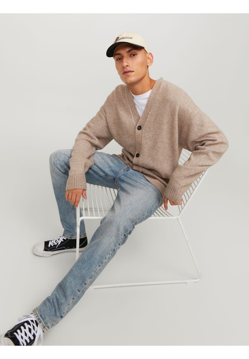 Cardigan beige lavorato a maglia con scollatura a V e tre bottoni neri, abbinato a jeans blu chiaro e sneakers nere, seduto su una sedia di rete.