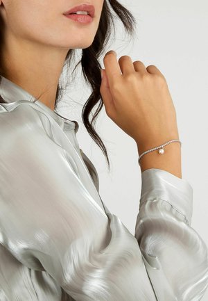Bracciale d'argento con link luccicanti e un singolo pendente in perla bianca, indossato su una mano contro una morbida blusa di seta grigia.