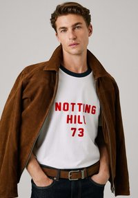 Καφέ σουέτ τζάκετ πάνω από ένα λευκό T-shirt με κόκκινο κείμενο "NOTTING HILL 73", σκούρο τζιν και καφέ ζώνη, με κλασικό σχέδιο.