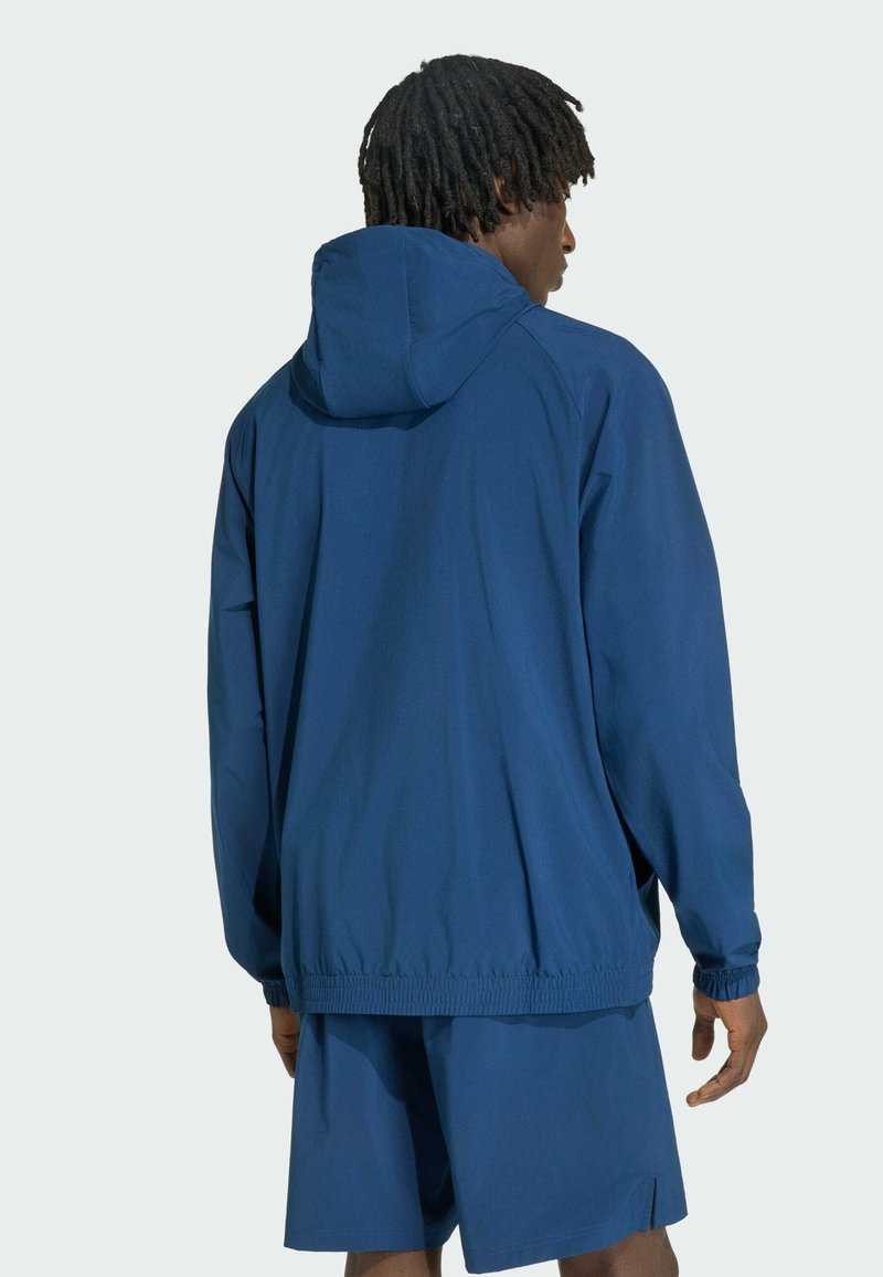 Mand med korte dreadlocks iført en blå hættetrøje og matchende blå shorts, vist bagfra mod en ensfarvet baggrund.