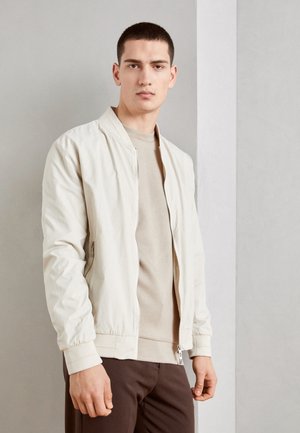 Giacca bomber beige chiaro con finitura testurizzata, polsini a costine e cerniera. Indossata sopra una maglietta beige semplice, abbinata a pantaloni scuri.